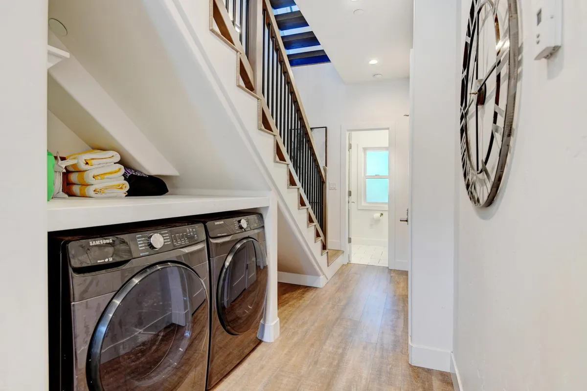 A functional yet welcoming laundry area with a full-size washer and dryer, staircase, and a clock on the wall. The space is bright and organized, suggesting convenience and ease for guests.