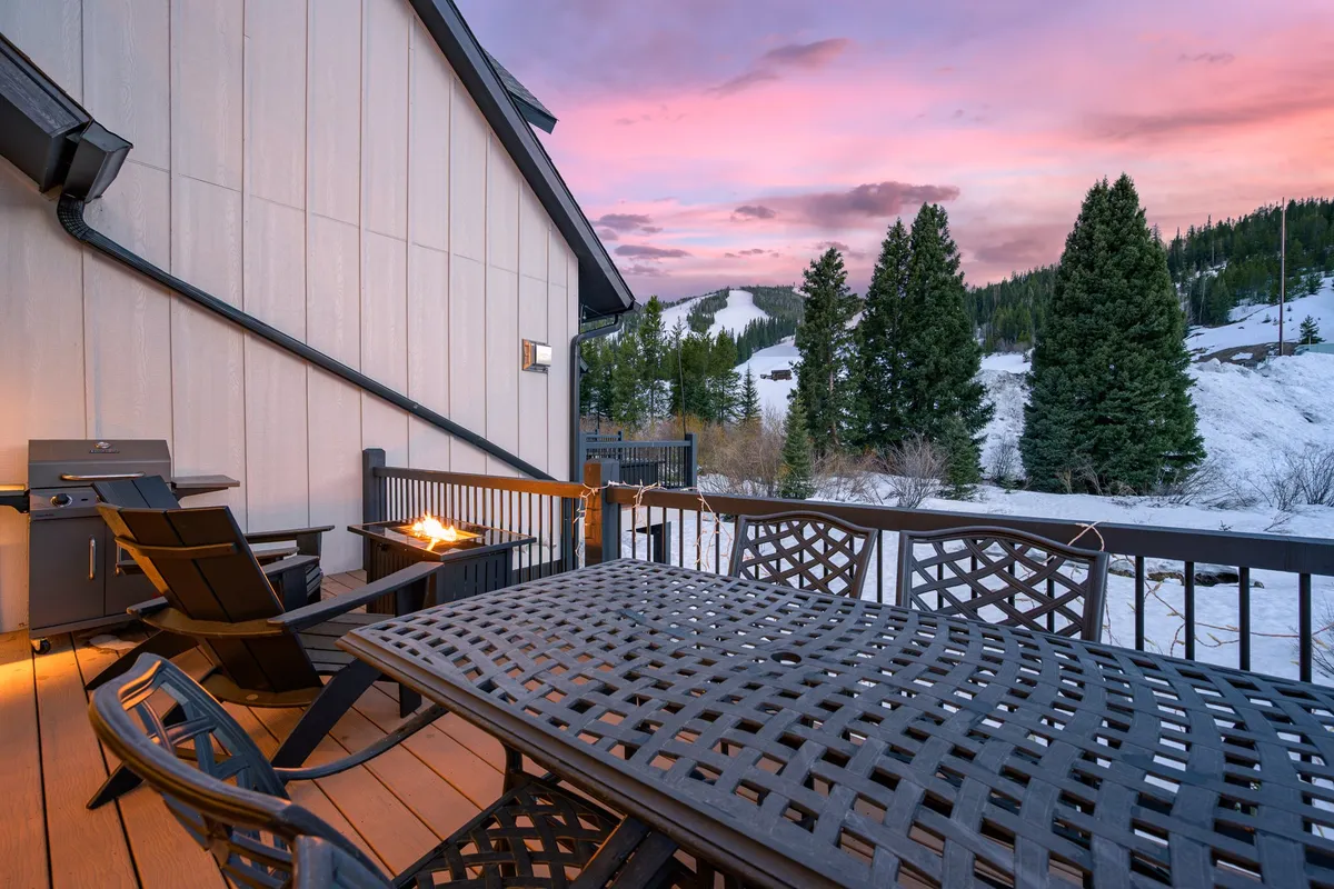A wooden upper deck set against a snow-covered landscape, featuring an outdoor dining table and adjoining BBQ grill. The deck provides views of the surrounding mountains and trees, ideal for outdoor feasts in all seasons.