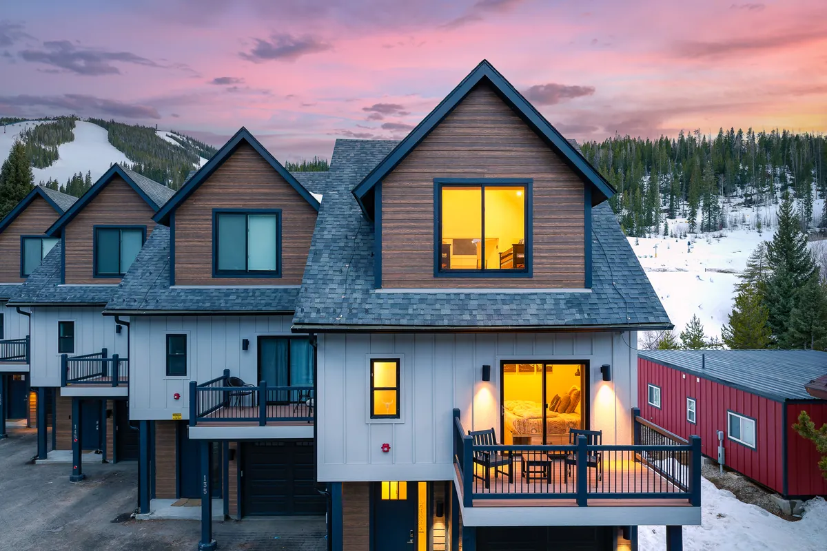 A modern architectural exterior with large windows and a deck furnished for relaxation. The backdrop showcases snowy slopes and a sunset sky, highlighting both winter and summer versatility for gatherings or quiet moments.