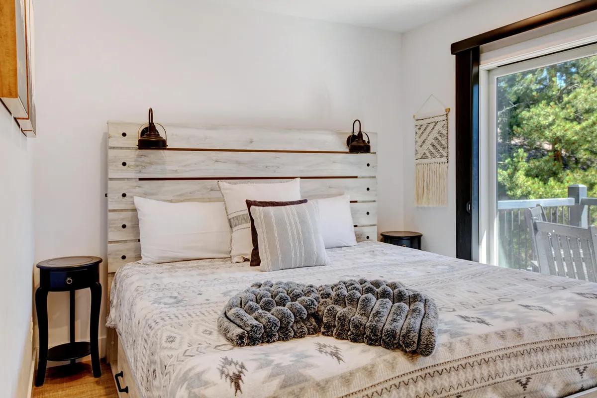 A bedroom featuring a wooden headboard, plush gray throw, decorative pillows, a nightstand with lamp, and a textured wall hanging. The scene exudes comfort and warmth, with natural light streaming in for a restful atmosphere.