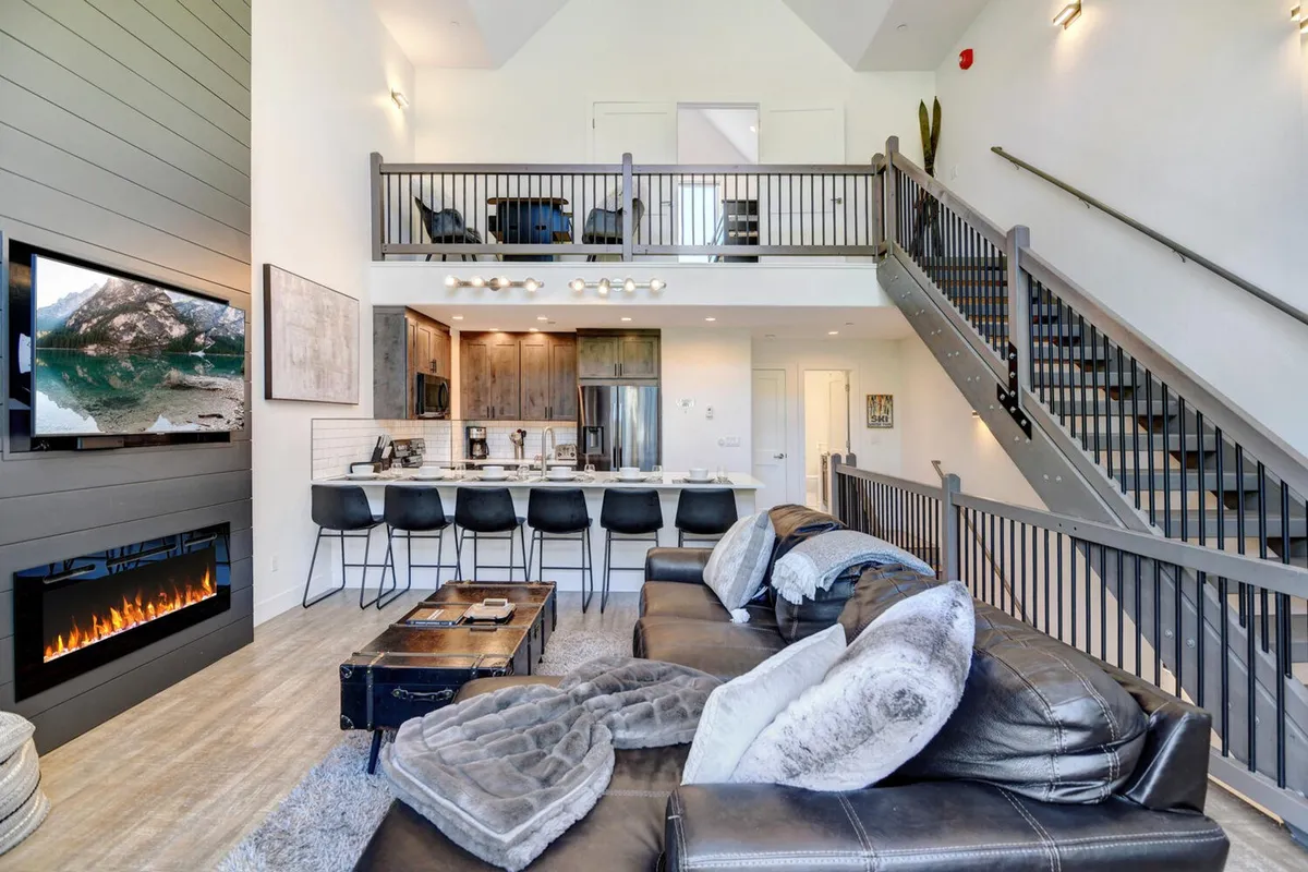 A modern great room with a comfortable sofa facing a fireplace and a large TV, adjacent to a kitchen island and stairs leading to upper levels. The space is designed for both entertainment and relaxation, with plenty of room for group activities.