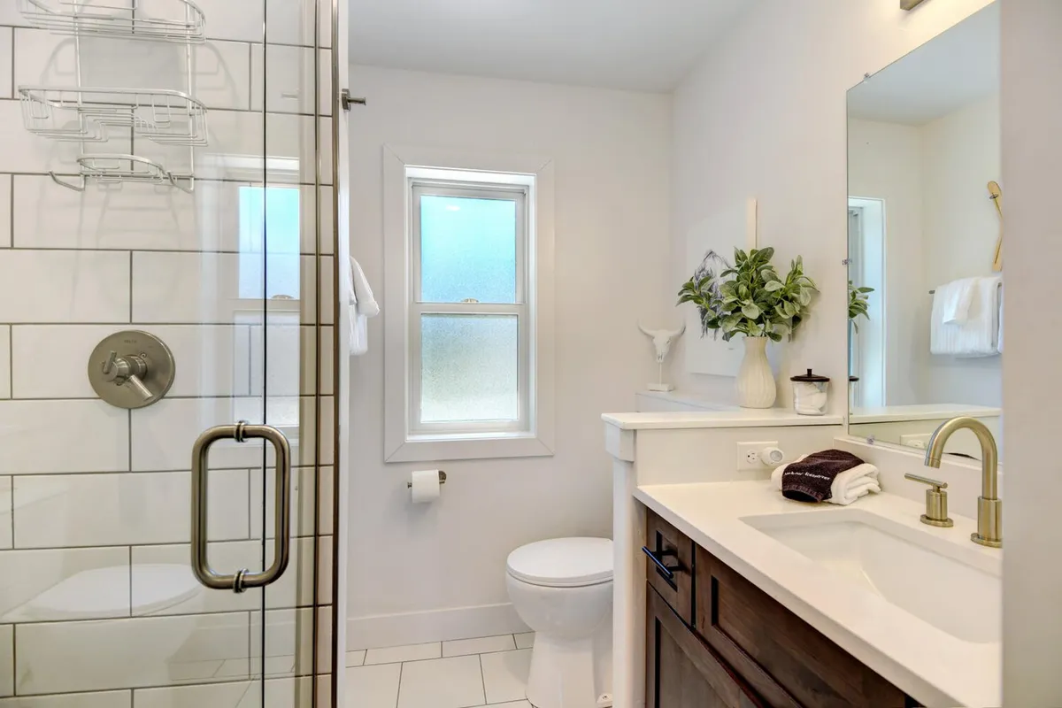 A calm, modern bathroom with a spacious shower area, vanity with sink, large window, and a decorative plant. The space feels clean, spa-like, and refreshing.