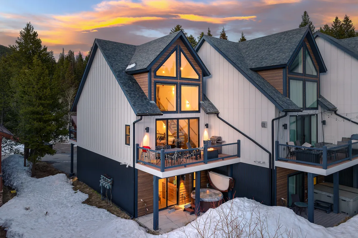 An inviting exterior view of the property with large windows glowing with warm indoor light, a balcony with comfortable seating, and a cozy fire pit area. The surrounding landscape hints at close proximity to nature and outdoor adventures.