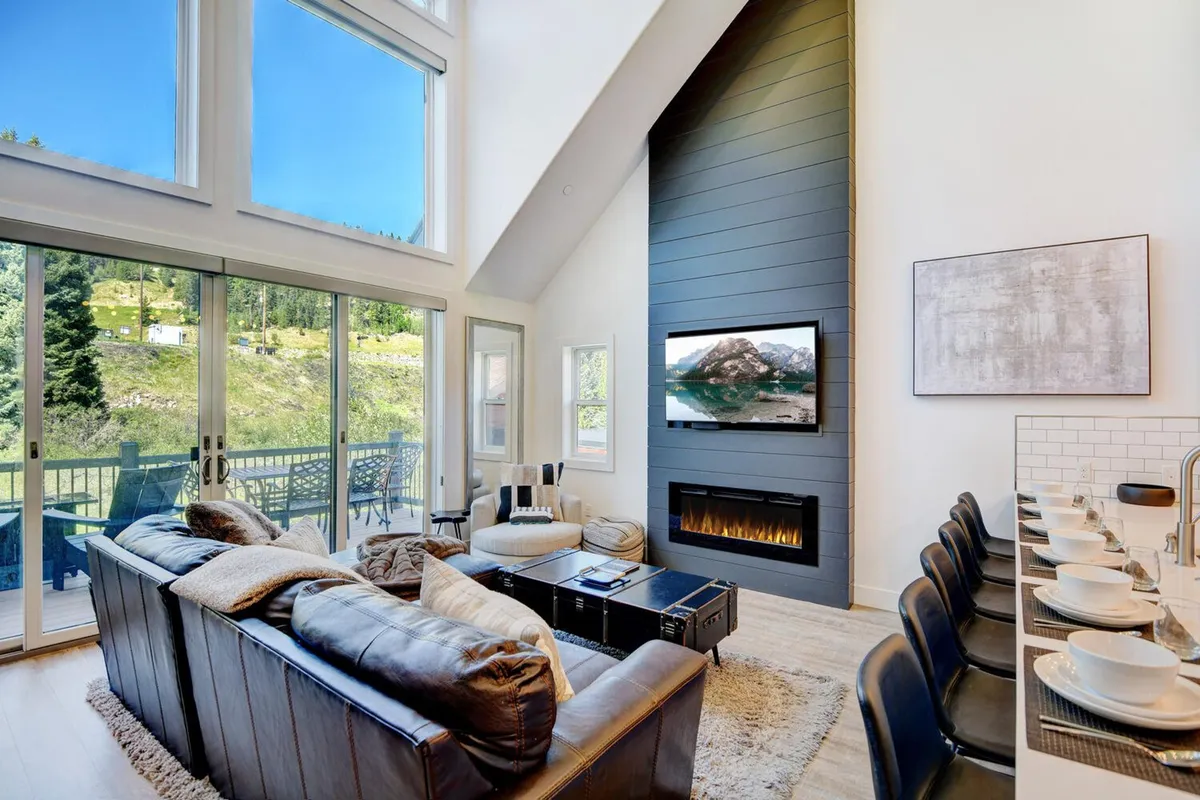 A welcoming great room with large windows framing mountain views, a modern fireplace set in a feature wall, plush and stylish seating, and a spacious dining area. Warm natural light fills the space, highlighting the seamless flow between relaxing and dining zones.