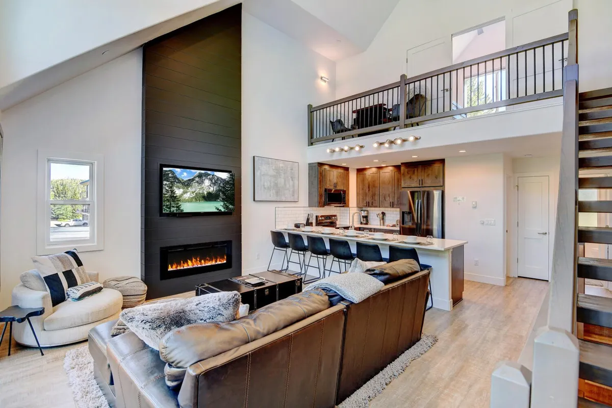 A spacious, open-concept living area featuring a large sofa, glowing fireplace, generous windows, and a kitchen island. The layout encourages relaxation, conversation, and shared moments, with natural light pouring in from multiple angles.