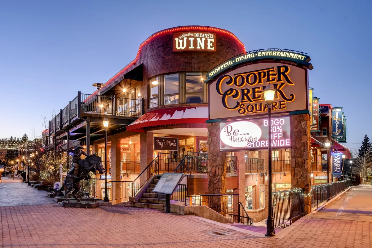A lively shot of Cooper Creek Square featuring the sign, wine shop, decorative lighting, and a statue. The festive atmosphere hints at shopping, dining, and entertainment opportunities in downtown Winter Park, a short shuttle ride away.