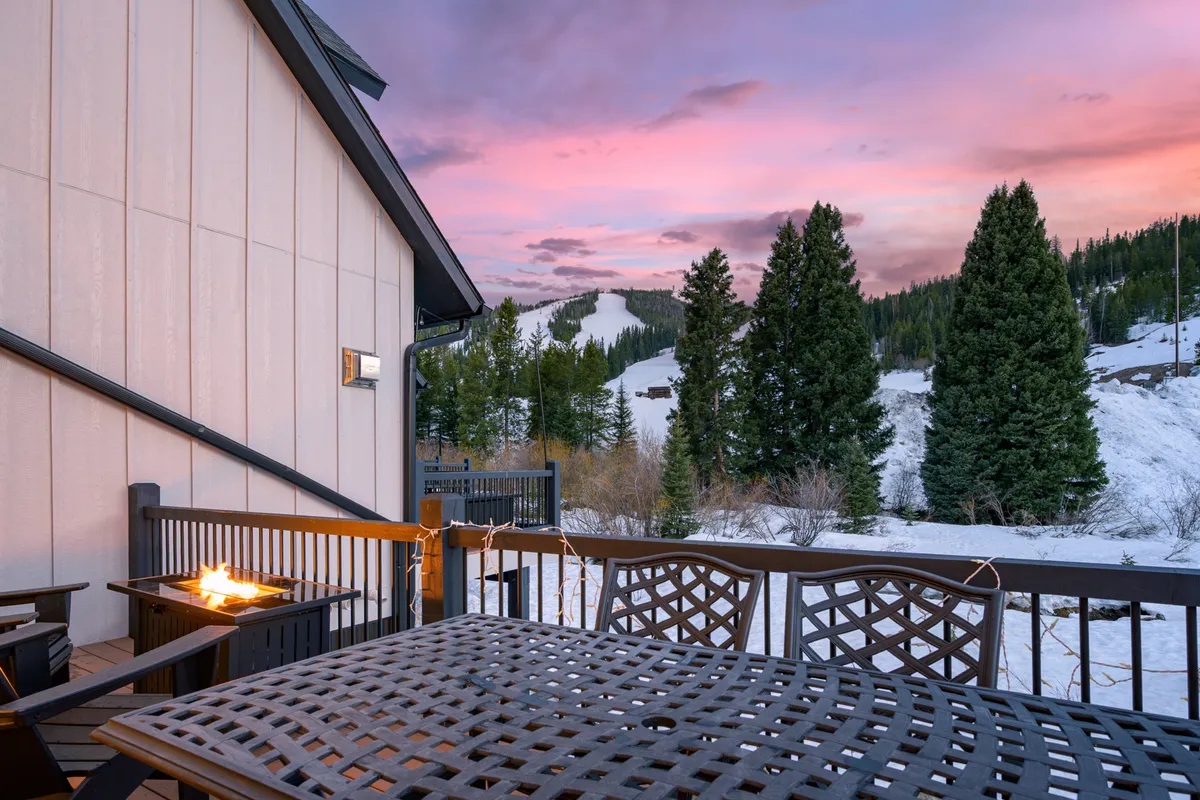 An exterior shot showcasing an outdoor dining table and fire pit with majestic mountain views and evergreen trees under a vibrant sunset sky. The setting invites relaxation and socializing after a day of adventure.